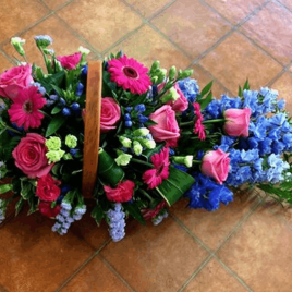 Bright and beautiful. A dark stained basket with flowers in the shape of a teardrop. Sky blue Delphiniums, Gentian, deep pink Roses, Cerise pink Gemini with a black eye and much more